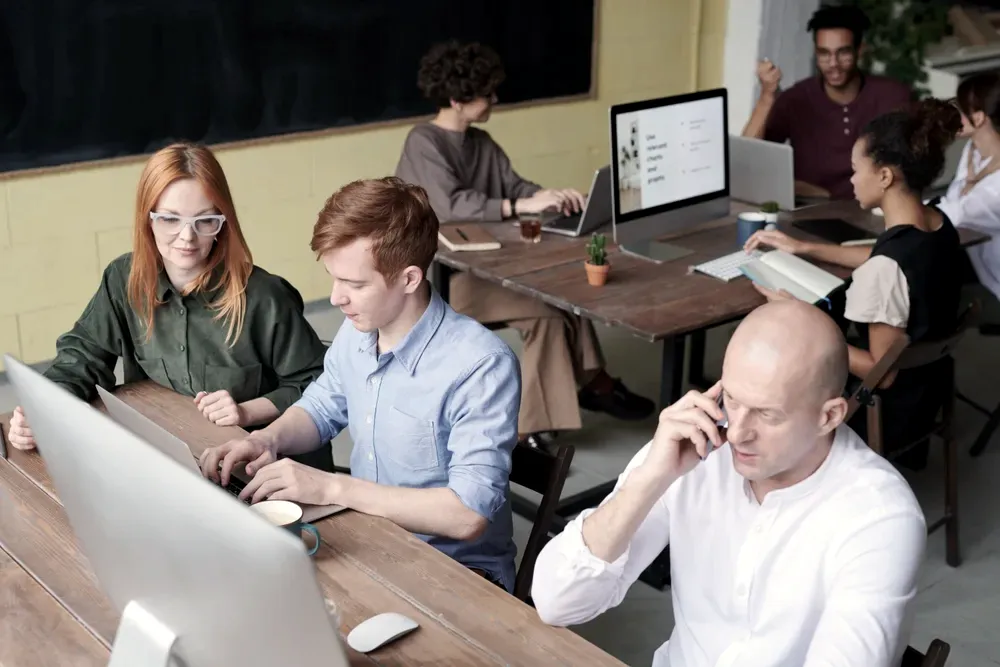 Open-plan office with several people working at shared wooden desks, one man on the phone in the foreground while others focus on their screens