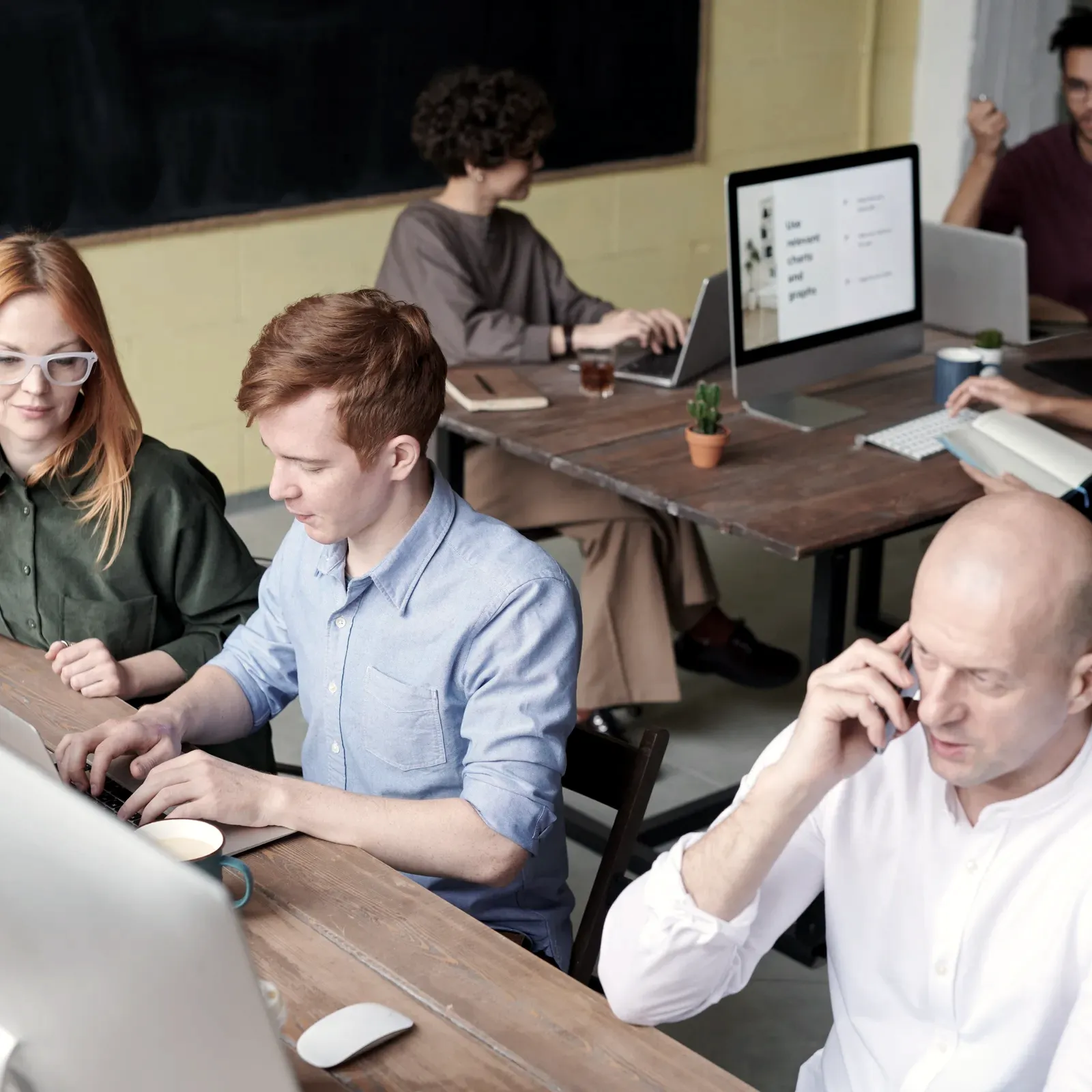 Open-plan office with several people working at shared wooden desks, one man on the phone in the foreground while others focus on their screens