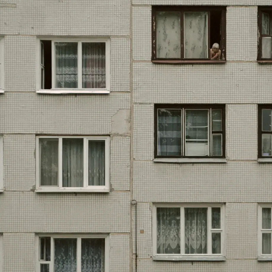 Tiled facade of an apartment block with net curtains in the windows and a resident looking out from an upper floor