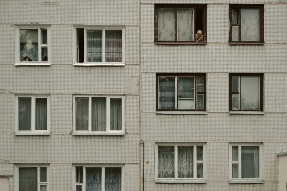 Tiled facade of an apartment block with net curtains in the windows and a resident looking out from an upper floor