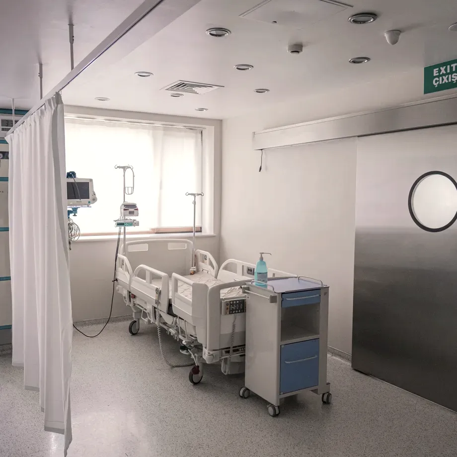 Empty modern hospital room with vital-signs monitors, IV stands and a steel sliding door, daylight from a window across the unoccupied bed.
