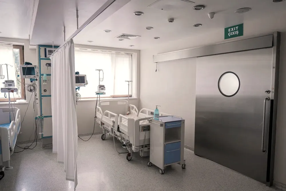 Empty modern hospital room with vital-signs monitors, IV stands and a steel sliding door, daylight from a window across the unoccupied bed.