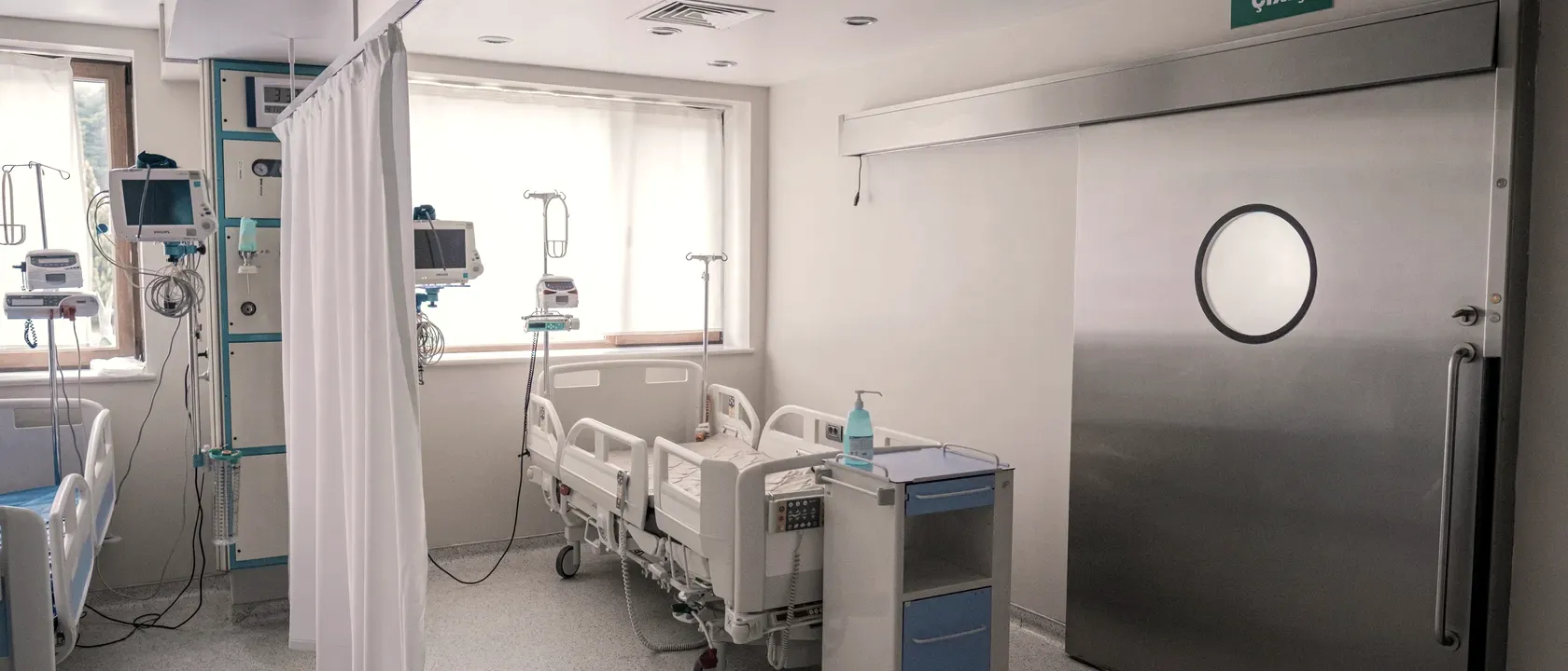 Empty modern hospital room with vital-signs monitors, IV stands and a steel sliding door, daylight from a window across the unoccupied bed.