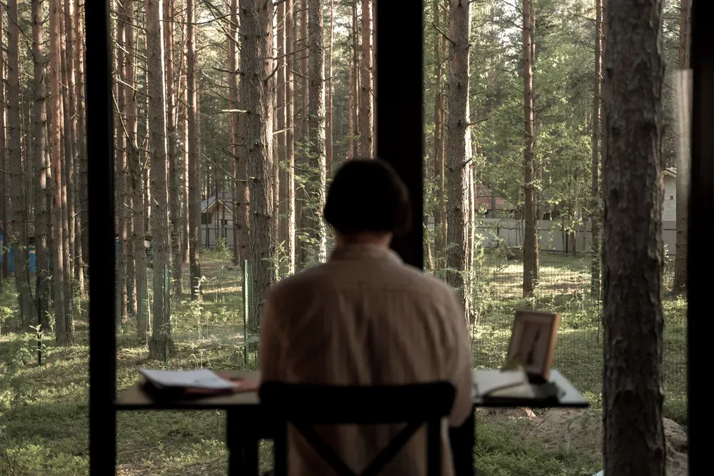 Person seen from behind working at a desk in front of a panoramic window overlooking a forest