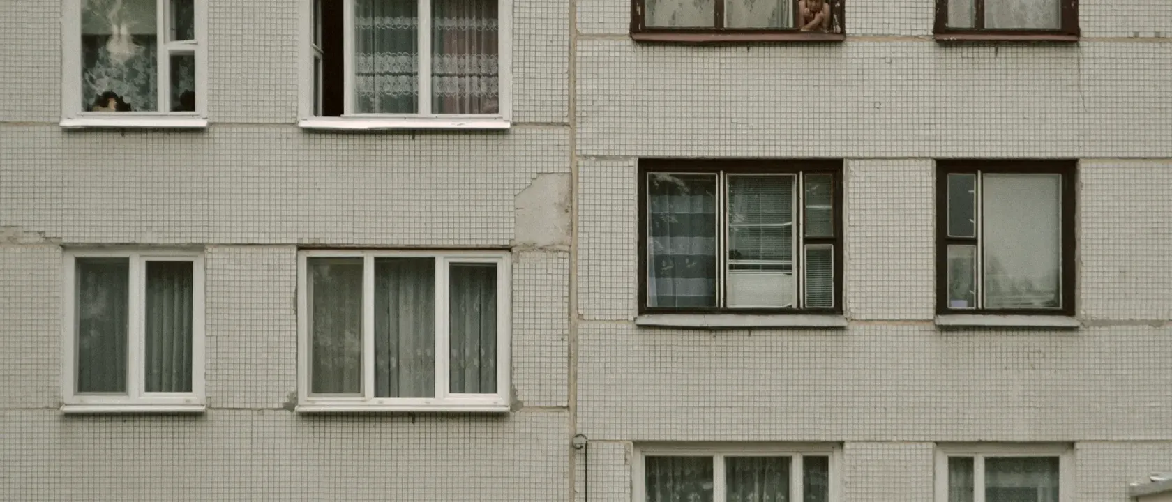 Tiled facade of an apartment block with net curtains in the windows and a resident looking out from an upper floor