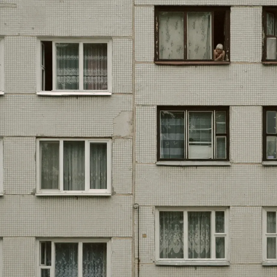 Tiled facade of an apartment block with net curtains in the windows and a resident looking out from an upper floor