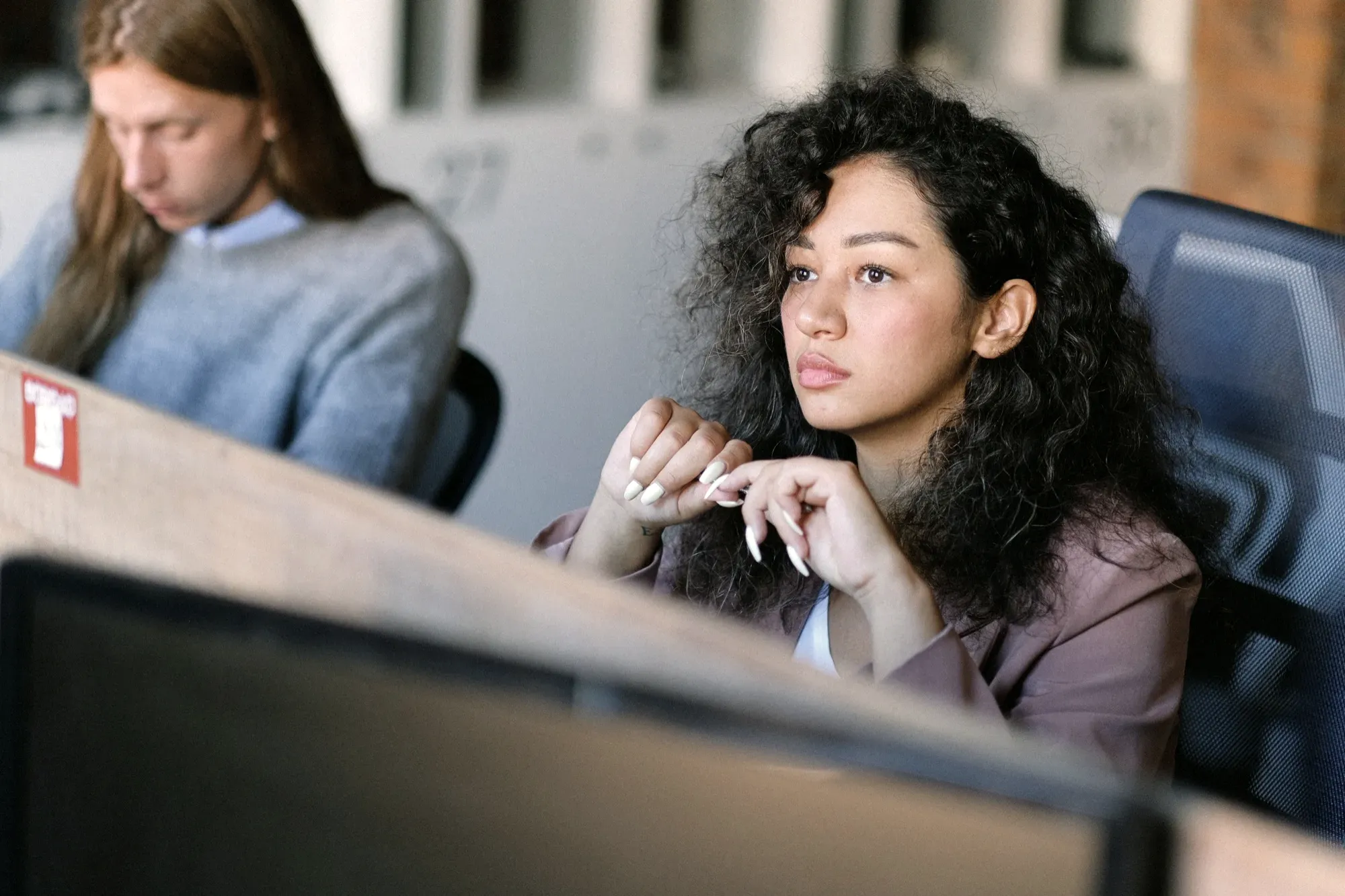 A person at a desk in an open-plan office, looking away from the screen in thought.